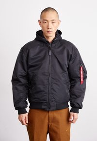 Black bomber jacket with a zip front, hood, ribbed cuffs, and hem. Features side pockets and a red fabric tag on the left sleeve.