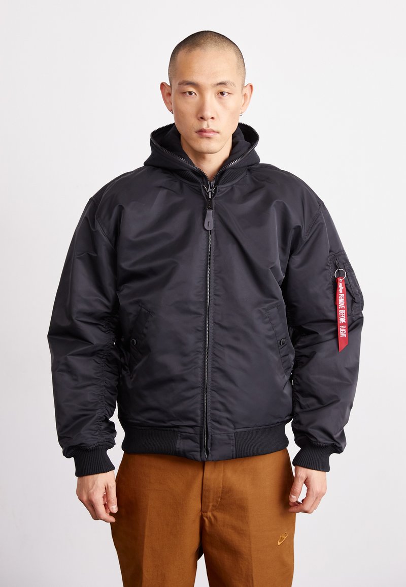 Black bomber jacket with a zip front, hood, ribbed cuffs, and hem. Features side pockets and a red fabric tag on the left sleeve.