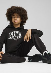 Fekete pamut kapucnis pulcsi "PUMA sports club" grafikával, hozzá illő jogger nadrág és fekete sportcipő zöld részletekkel. A modell ül, és az egyik karját a térdére támasztja.