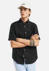 Schwarzes Kurzarm-Hemd mit Knopfleiste und Brusttasche, aus einem strukturierten Stoff; das Modell trägt eine weiße Mütze mit schwarzem Text.