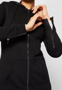 Hoodie noir à fermeture éclair avec capuche, deux poches avant et détail zippé sur la manche. Confectionné en tissu doux, avec des poignets texturés et une fermeture éclair.
