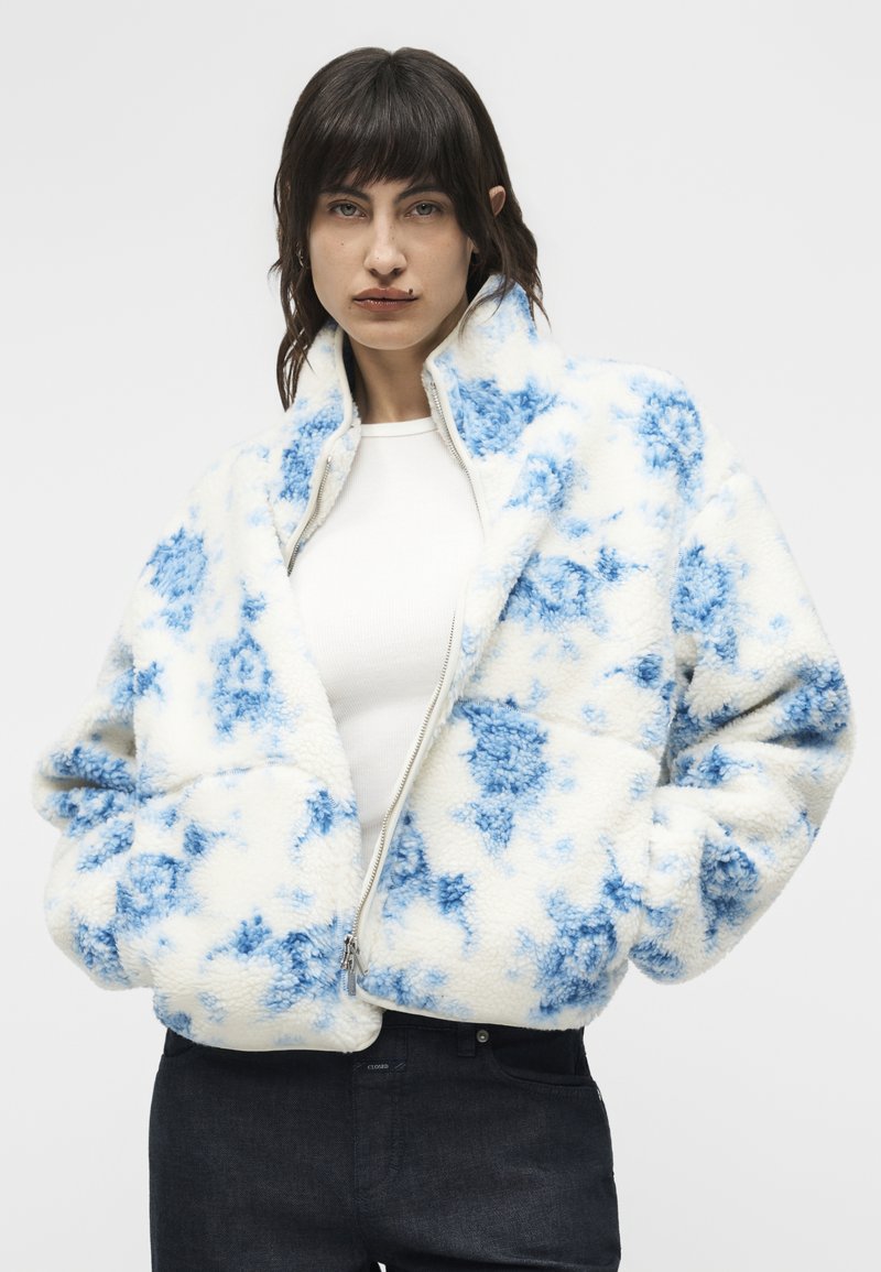 Frau trägt eine weiße und blaue gemusterte, flauschige Jacke mit Reißverschluss über einem weißen Hemd und dunkler Hose, steht mit den Händen in den Taschen.