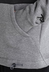 Šedá mikina s kapsou, stahovací šňůrkou na lemu a černým logem na rukávu. Měkká textura a sportovní design.