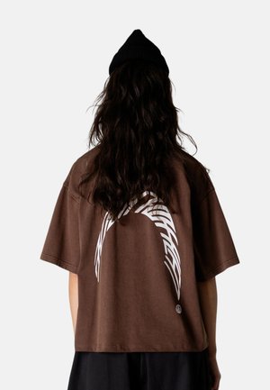 Person med langt mørkt hår og sort beanie har en stor brun T-shirt med hvidt abstrakt buedesign på ryggen.