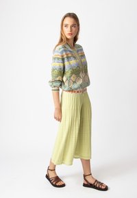 Žalios megztos bomber tipo striukės su daugiaspalviais raštais komplektas su šviesiai žalia raukinėta sijonu ir rudais paveikslinio dirželio sandalais.