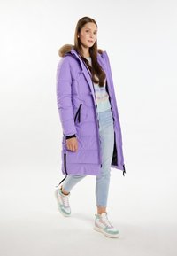 Pardesiu lung pufos de culoare mov, cu glugă detașabilă din blană artificială, buzunare laterale și accente negre. Asortat cu blugi bleu deschis și adidași multicolori.