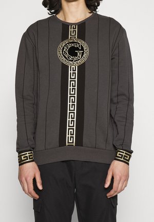 Mann trägt dunkelgrauen Pullover mit vertikalem schwarzem und goldenem griechischem Schlüssel-Muster und rundem Emblem mit einem "G" auf der Brust.