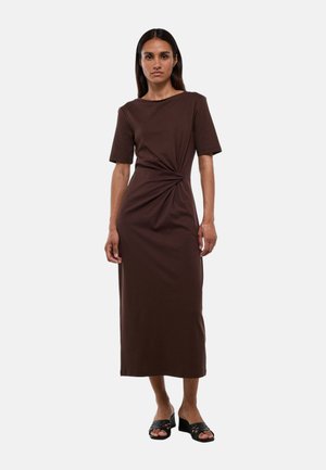 Femme debout face à l'avant portant une robe midi marron à manches courtes avec un détail torsadé sur le côté, associée à des sandales à talons ouvertes noires.