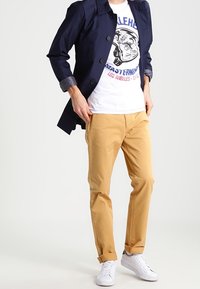 Granatowy wodoodporny płaszcz przeciwdeszczowy, biały t-shirt z grafiką, beżowe chinosy i białe trampki; guziki i podwinięte rękawy na płaszczu.