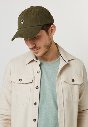 Man met een olijfgroene corduroy pet met boomlogo, een lichtbeige jasje met knopen over een groene ronde hals T-shirt, kijkend naar beneden.