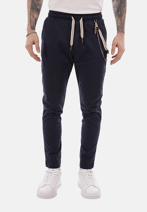 Uomo che indossa pantaloni jogger blu navy con coulisse bianche e accessorio a catena, abbinati a sneaker bianche e una camicia bianca.