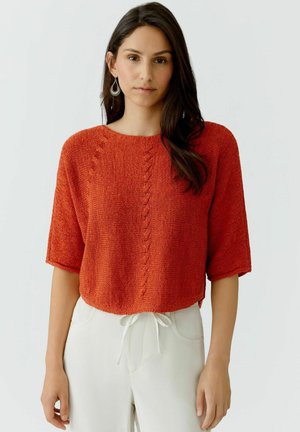 Vrouw met lang donker haar, zilveren oorbellen in de vorm van ringen, een oranje gebreide trui met korte mouwen en witte broek met een aantrekkoord in de taille.