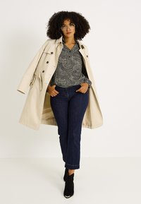 Abrigo beige estilo gabardina, blusa negra con estampado, vaqueros azul oscuro de pierna recta y botines negros. Pelo rizado y gafas completan el conjunto.