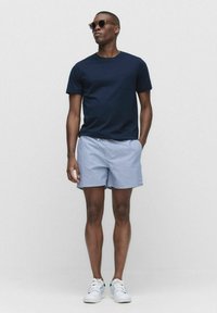 Marineblauwe T-shirt met korte mouwen en zak, gecombineerd met gestreepte blauwe shorts. Model draagt een zonnebril en witte sneakers, staande tegen een neutrale achtergrond.