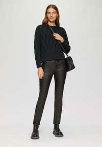 Donna in un maglione a maglia cablata nero, pantaloni di pelle neri, stivali neri e borsa a tracolla nera, in piedi contro uno sfondo chiaro.