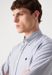 Lichtblauw overhemden van zachte stof, met een button-down kraag en een klein zwart logo op de borst. Glad textuur.