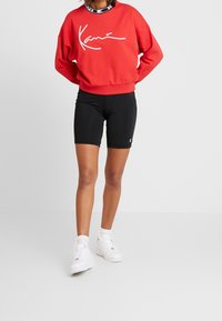 Sudadera roja con logo de firma en blanco, combinada con pantalones cortos de ciclista negros. El modelo lleva zapatillas y calcetines blancos, de pie sobre un fondo claro.