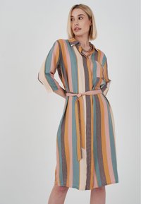 Frau in einem knielangen, kragenlosen Kleid mit vertikalen Streifen in Senfgelb, Türkis, Rosa, Creme und Braun, mit einem Gürtel an der Taille gebunden.