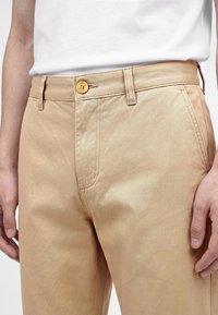 Pantalon beige avec passants de ceinture et fermeture par bouton, porté par une personne avec les mains détendues le long du corps.