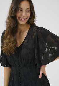 Abito nero realizzato in tessuto trasparente con texture floreale. Presenta maniche corte e fluide e una scollatura a V con bottoni sulla parte anteriore.