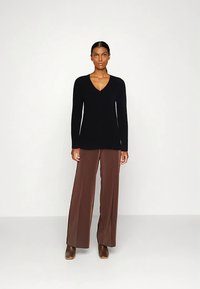 Zwart V-hals sweater gemaakt van zacht materiaal, gecombineerd met brede bruine broek. Model staat met neutrale uitdrukking tegen een effen achtergrond.