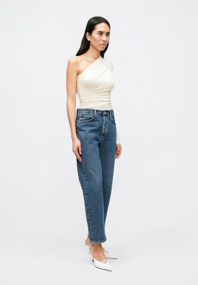 Top branco de um ombro só com textura franzida, combinado com jeans azuis de cintura alta. A modelo usa sapatos de salto branco com bico pointed, em frente a um fundo neutro.