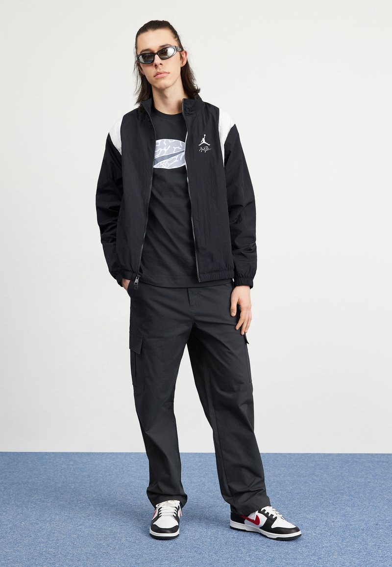 Veste zippée noire avec des accents blancs, t-shirt graphique noir, pantalon cargo, et baskets noir et blanc avec des détails rouges.