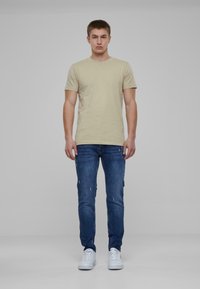 Bež pamukasta majica, slim-fit kroja, uparena s plavim iznošenim trapericama i bijelim tenisicama. Model stoji uspravno protiv neutralne pozadine.