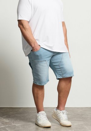 Mężczyzna w białych trampkach, jasnoniebieskich dżinsowych szortach do kolan i białej koszuli z krótkim rękawem, stoi z rękami w kieszeniach.