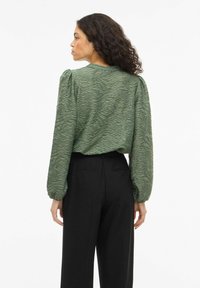 Blusa verde con textura y mangas largas abullonadas, combinada con pantalones negros de talle alto. La blusa presenta un patrón de ondas en la tela.