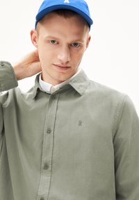 Olijfgroene button-up shirt met een gestructureerde afwerking, voorzien van een klein geborduurd logo. Draagt een blauwe pet, wat een casual stijl laat zien.