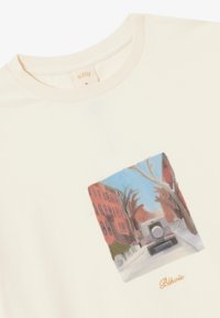 Cremfarbener Baumwoll-T-Shirt mit einem quadratischen Grafikmotiv einer Straßenszene, die rote Gebäude und ein Fahrzeug zeigt, mit "Blkwvs" in Gold gestickt.