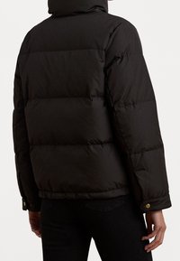 Henkilö, joka on pukeutunut mustaan vuorattuun toppatakkille, jossa on huppu, näkyy takaa päin, yllään mustat housut.