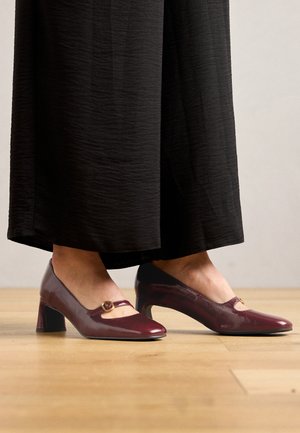 Burgundy kiilto nahkakengät, joissa on pyöreä kärki, Mary Jane -varsi ja pieni kultainen yksityiskohta, teksturoidulla puulattialla.