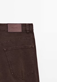 Pantalon en denim marron foncé avec une poche arrière et une étiquette en cuir marron cousue à la ceinture.