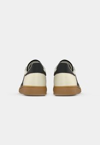 Pareja de zapatillas de deportivo de caña baja con parte superior beige, detalles en negro en el talón y suelas de goma vista desde la parte trasera sobre un fondo blanco.