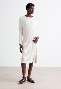 Knit midi-mekko vaaleassa sävyssä, jossa on pystysuora ribbitekstuuri, pitkät hihat ja pyöreä pääntie. Yhdistetty mustiin teräväkärkisiin kenkiin, joissa on niittejä.