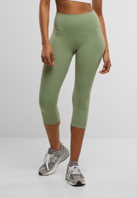 Groene capri leggings van rekbaar materiaal, met een hoge tailleband en een gladde textuur. Gecombineerd met grijze sportschoenen.
