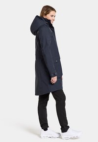 Navy lange jas met capuchon, voorzak en ritssluitingen. Gecombineerd met zwarte broeken en witte sneakers. Gladde textuur en getailleerde pasvorm.