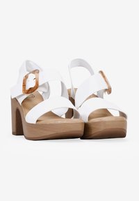 Sandales à talons hauts blanches avec de larges lanières et une boucle couleur beige, présentant une semelle en plateforme texturée en bois et une finition en cuir lisse.