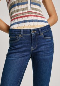 Kvinna som bär blå denimjeans och en ärmlös ribbad stickad topp med horisontella ränder i röd, blå, beige och vit.