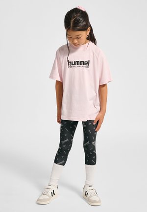 Jong meisje staat, draagt een roze oversized "hummel performance" t-shirt, zwarte patterned leggings, witte sokken en witte sneakers.