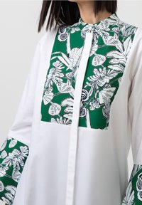 Camicia bianca con pannelli a motivo floreale verde, caratterizzata da un colletto alto, chiusura a bottoni e maniche lunghe con dettagli abbinati. Tessuto liscio.