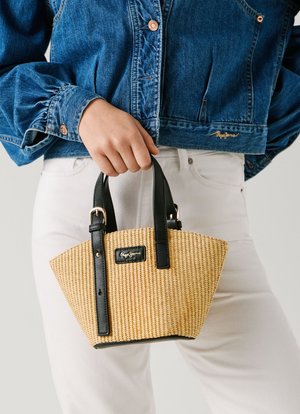 Osoba u bijelim hlačama i plavoj traper jakni držeći malu bež pletenu torbicu s crnim ručkama i detaljem kopče.