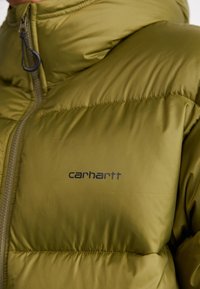 Giacca isolante verde oliva con texture trapuntata. Presenta una cerniera intera, colletto alto e logo Carhartt sul petto.