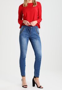 Žena oblečená v jasně červeném dlouhém rukávu, modrých těsně přiléhajících džínách s nastavitelným pasem a černých sandálech na vysokém podpatku, stojící na bílém pozadí.