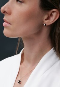 Collana con pendente in tonalità oro con una pietra nera rotonda, abbinata a piccoli orecchini a bottone neri. Texture liscia su uno sfondo neutro.