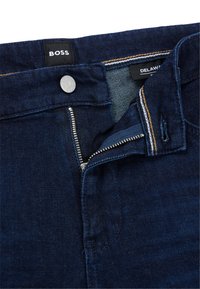 Jean en denim bleu foncé avec braguette zippée, fermeture par bouton métal et surpiqûres apparentes en couleurs contrastantes. Étiquette de la marque visible.