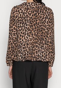 Bluusi, jossa on leopardikuvio, peplum-muotoilu, pitkät hihat ja röyhelökaulus. Valmistettu pehmeästä kankaasta ruskean ja mustan sävyissä.