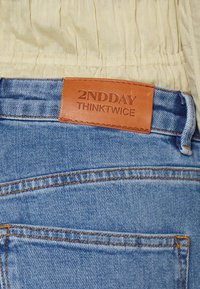 Jeans de mezclilla azul claro con costuras naranjas y una etiqueta de cuero marrón con la inscripción "2NDDAY THINKTWICE" en la cinturilla.